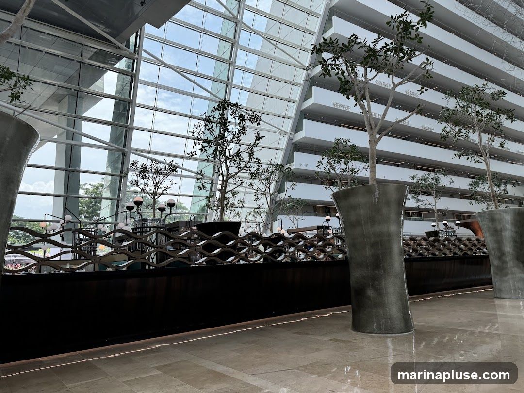 The image shows the interior of a large, modern building with a high ceiling and a glass facade. Several potted trees are placed throughout the space, adding a touch of nature to the architectural design. The floors are tiled, and the overall ambiance is one of spaciousness and contemporary elegance.