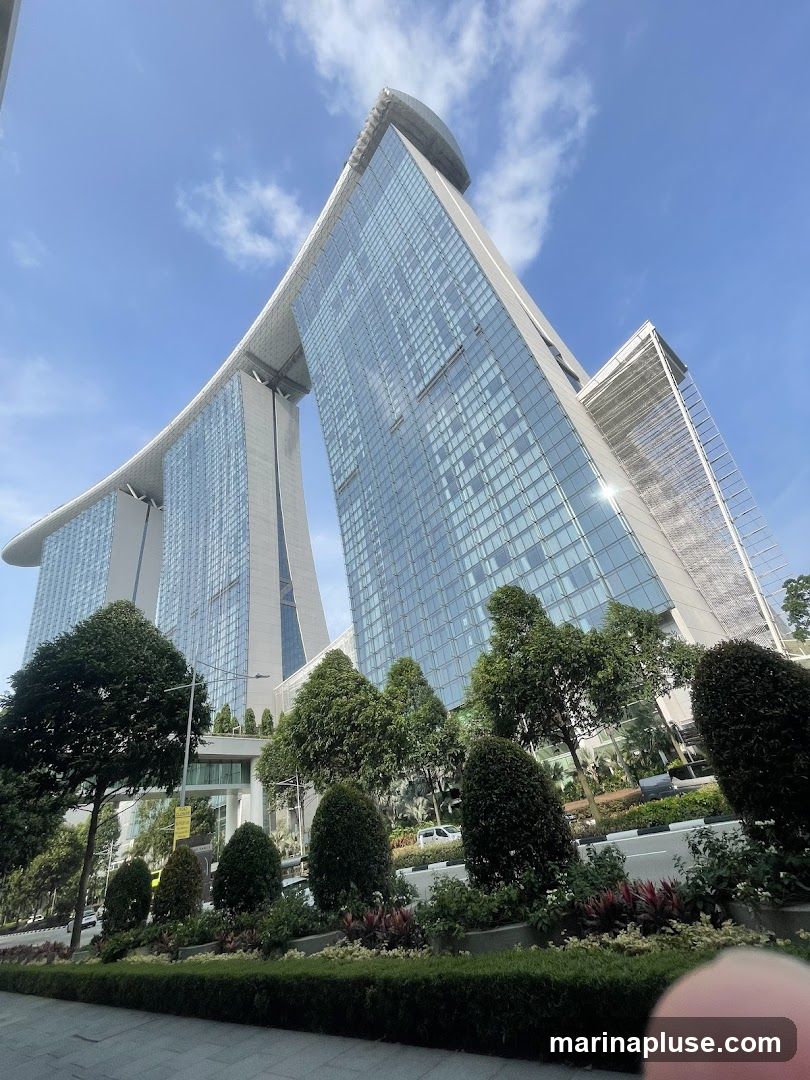 A low-angle shot of the Marina Bay Sands hotel in Singapore on a clear day. The iconic building, with its three towers topped by a large, boat-like structure, is seen against a bright blue sky with scattered clouds. Lush green trees and manicured bushes line the foreground, with a sidewalk and a road visible.