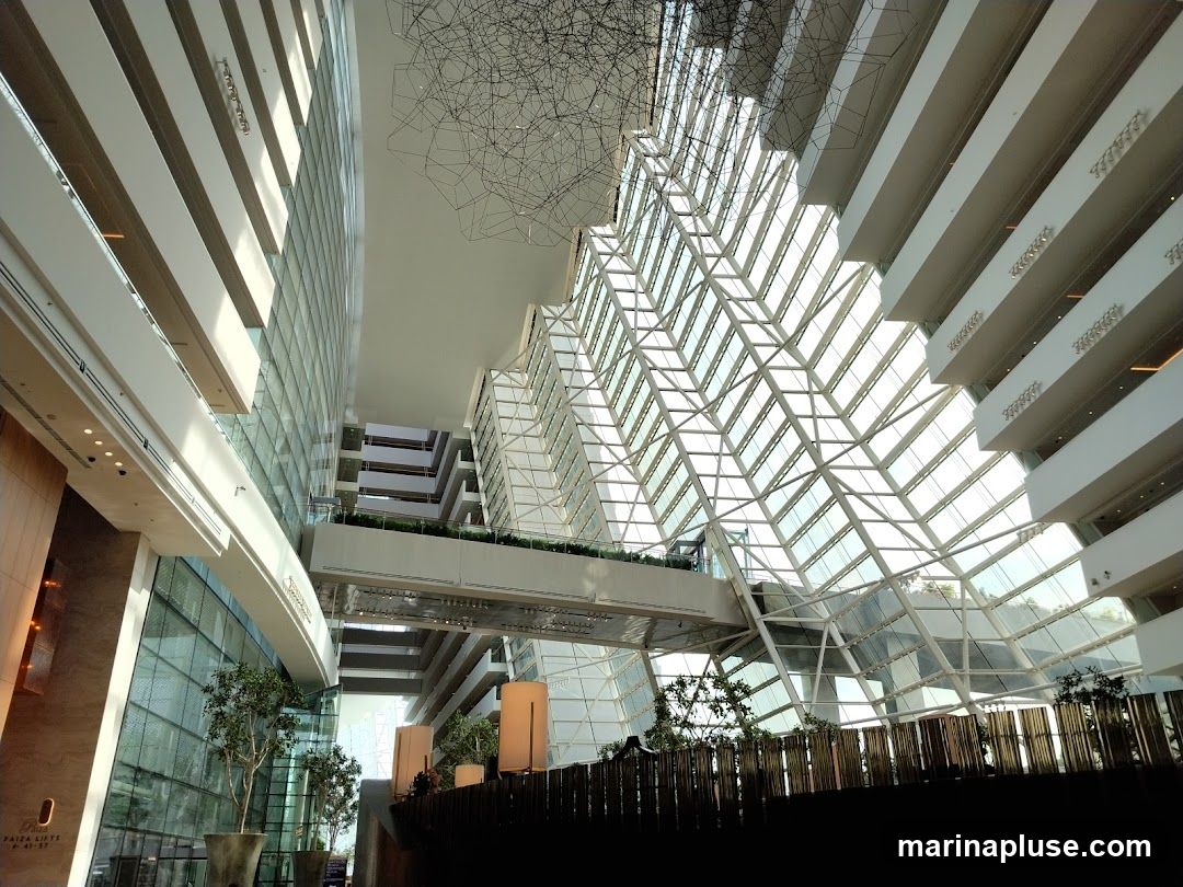 A wide-angle shot of a grand, modern atrium with a striking geometric glass ceiling. Sunlight streams through the angled panes, illuminating the multi-level interior. Balconies with white railings overlook the central space, and a complex wire sculpture hangs from the ceiling. Lush potted plants are visible on the lower levels, adding a touch of nature to the architectural design.