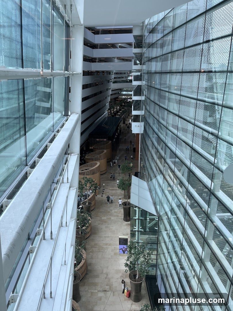 An interior view of a multi-story modern building with a central atrium. Large glass walls line the sides, reflecting the interior. Several levels of walkways and balconies are visible, with people walking below. Decorative wooden planters with trees and plants are placed along the walkways. The floor is paved with light-colored tiles.