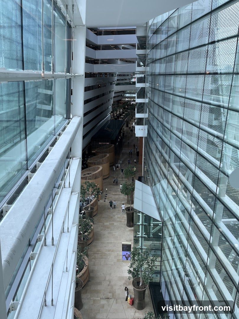 An interior view of a multi-story modern building with a central atrium. Large glass walls line the sides, reflecting the interior. Several levels of walkways and balconies are visible, with people walking below. Decorative wooden planters with trees and plants are placed along the walkways. The floor is paved with light-colored tiles.