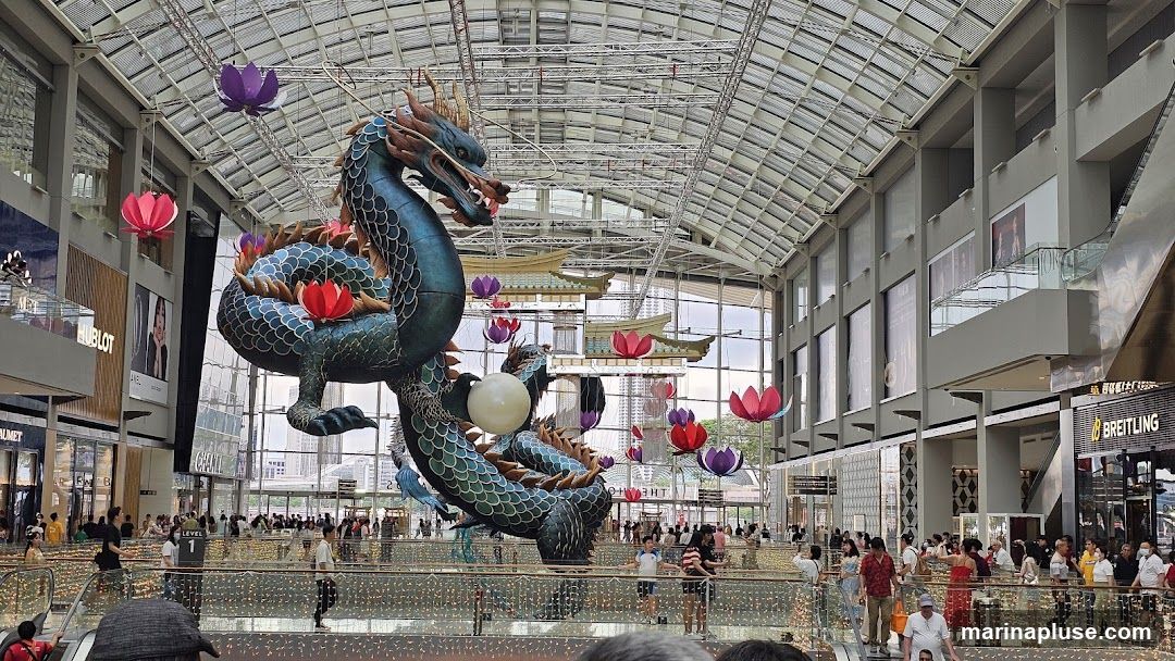 A large, ornate dragon sculpture hangs from the ceiling of a modern shopping mall, adorned with lotus flower decorations. The mall features multiple levels with escalators and shops, including visible signage for brands like HUBLOT and BREITLING. People are seen browsing and walking around the mall.