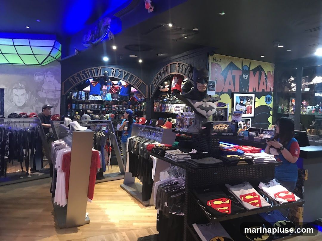 Interior view of a store selling merchandise related to comic book characters, primarily Batman and Superman. Racks of t-shirts with character logos are displayed, along with hats and other apparel. A bust of Batman is prominently featured, along with posters and artwork on the walls. Two staff members are visible assisting customers.
