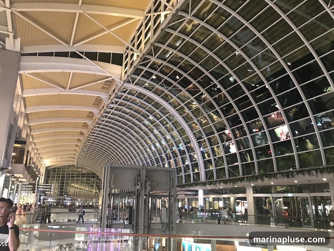 A wide-angle view of a modern interior space with a distinctive arched glass ceiling and a white, geometric support structure. The ceiling reflects the lights and activity of the space below, including glimpses of shops and people. The overall impression is one of a large, contemporary shopping mall or airport terminal.
