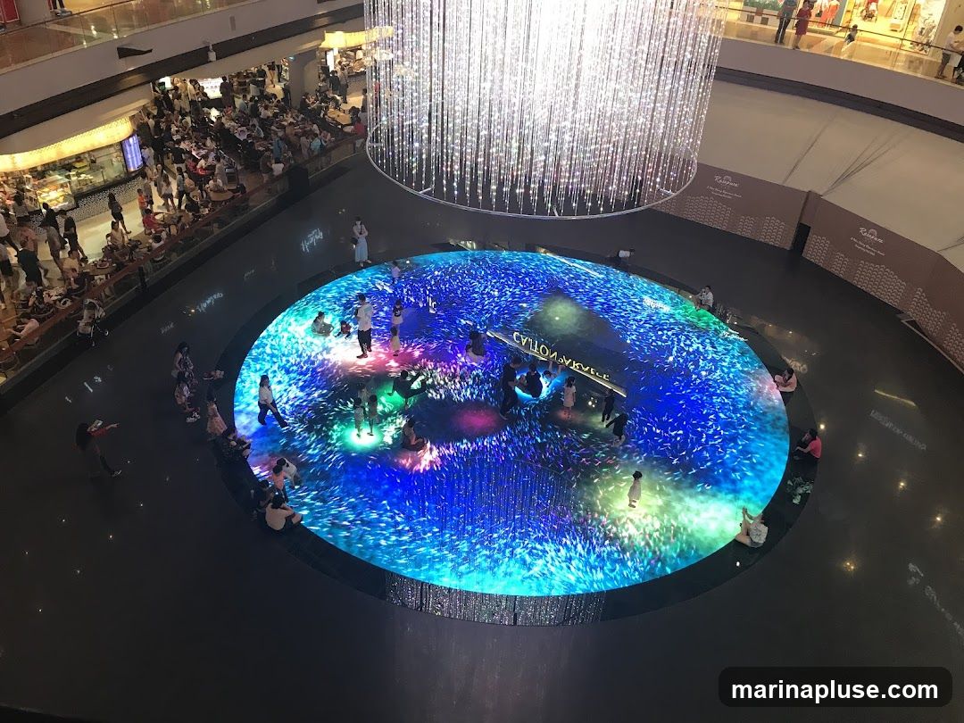 An overhead view of a busy shopping mall atrium. In the center, a large circular LED display shows a vibrant, animated underwater scene with schools of fish. People are gathered around the display, some sitting and watching, while others stand and interact with the projections. Above the display, a massive, sparkling chandelier hangs, casting a bright light. In the background, multiple levels of the mall are visible, with shops and restaurants, and many shoppers walking around.