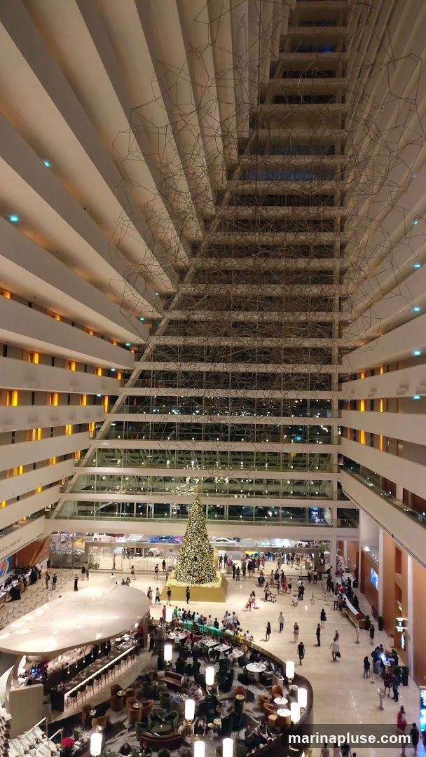 An aerial view of a large, multi-story hotel atrium decorated for Christmas. A tall, ornate Christmas tree stands in the center of the ground floor, surrounded by people. Several levels of balconies overlook the atrium, with lights illuminating the interior. A modern bar and seating area are visible in the foreground. Abstract wire sculptures hang from the ceiling.