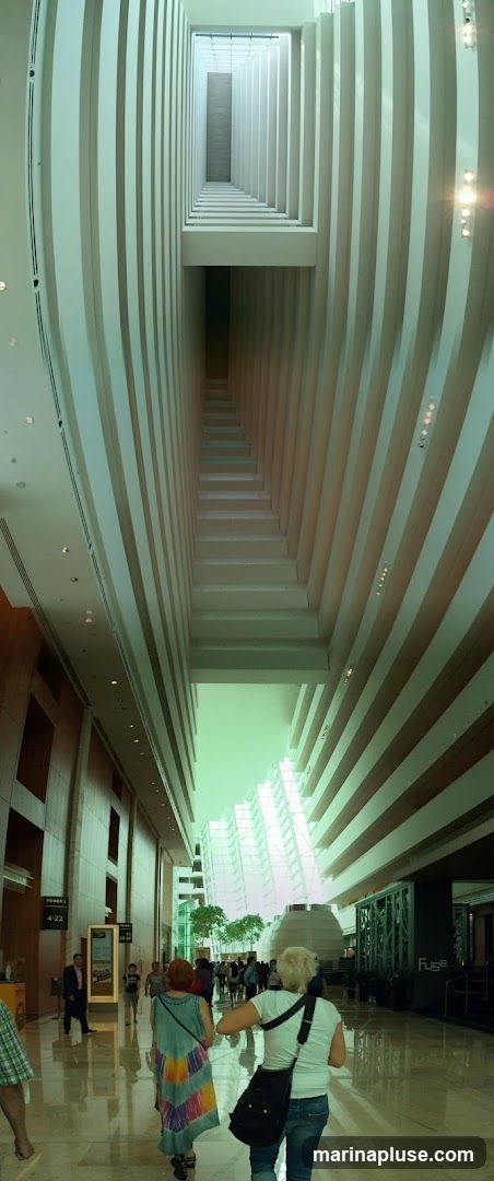 A panoramic view looking up through a modern, multi-story atrium. The architecture features repeating vertical fins that create a sense of depth and perspective, drawing the eye upwards towards a skylight. People are seen walking on the polished floor below, with some trees and modern building structures visible in the distance through the open space.