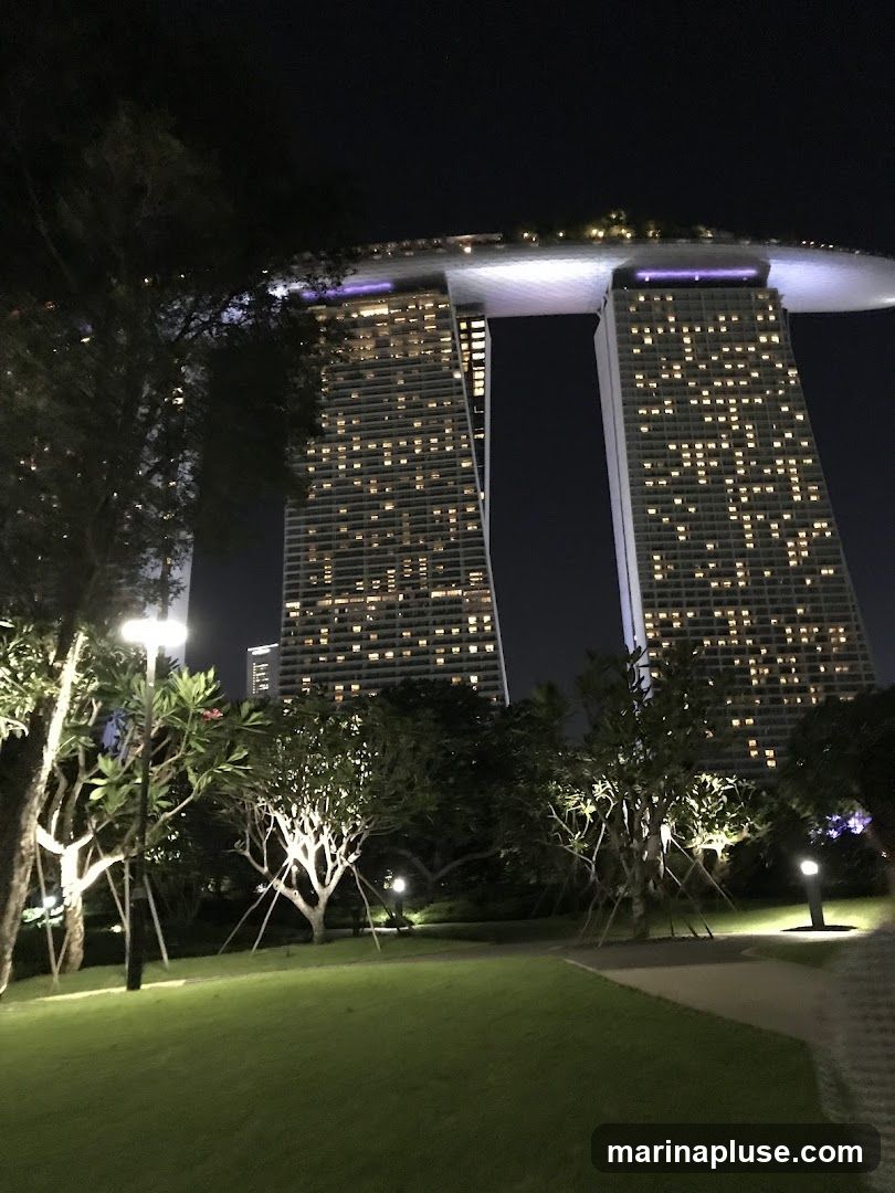 A nighttime view of the Marina Bay Sands hotel in Singapore, with its iconic three towers and connecting SkyPark. The hotel is illuminated, and the surrounding gardens are lit by lampposts.