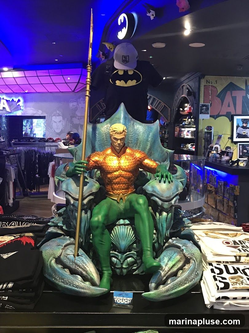 A large statue of Aquaman seated on a throne resembling a crab is displayed in a store. He is holding a trident. The statue is surrounded by merchandise, including t-shirts with various designs, some featuring the Batman logo. In the background, shelves are stocked with more merchandise, and Batman-themed decor is visible on the walls and ceiling. A sign that reads "PLEASE DO NOT TOUCH" is placed near the base of the statue.