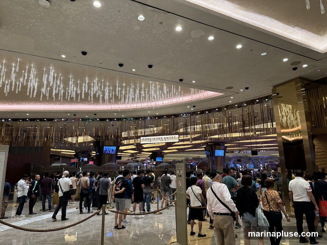 A large group of people are standing in line inside a brightly lit, modern casino. The ceiling features decorative lighting fixtures, and the walls are adorned with metallic accents. A sign indicates 'FOREIGNER ENTRY'.