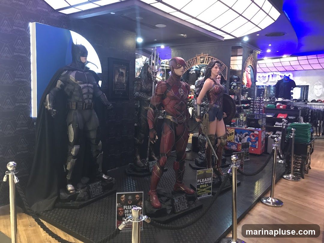 A retail display featuring life-sized figures of Batman, The Flash, and Wonder Woman from the Justice League. The figures are positioned on a black textured platform with "JUSTICE LEAGUE" signage. In the background, shelves are stocked with merchandise, including t-shirts and other apparel, with a blue and black ceiling overhead. Signs with "PLEASE DO NOT ENTER" are visible near the figures.