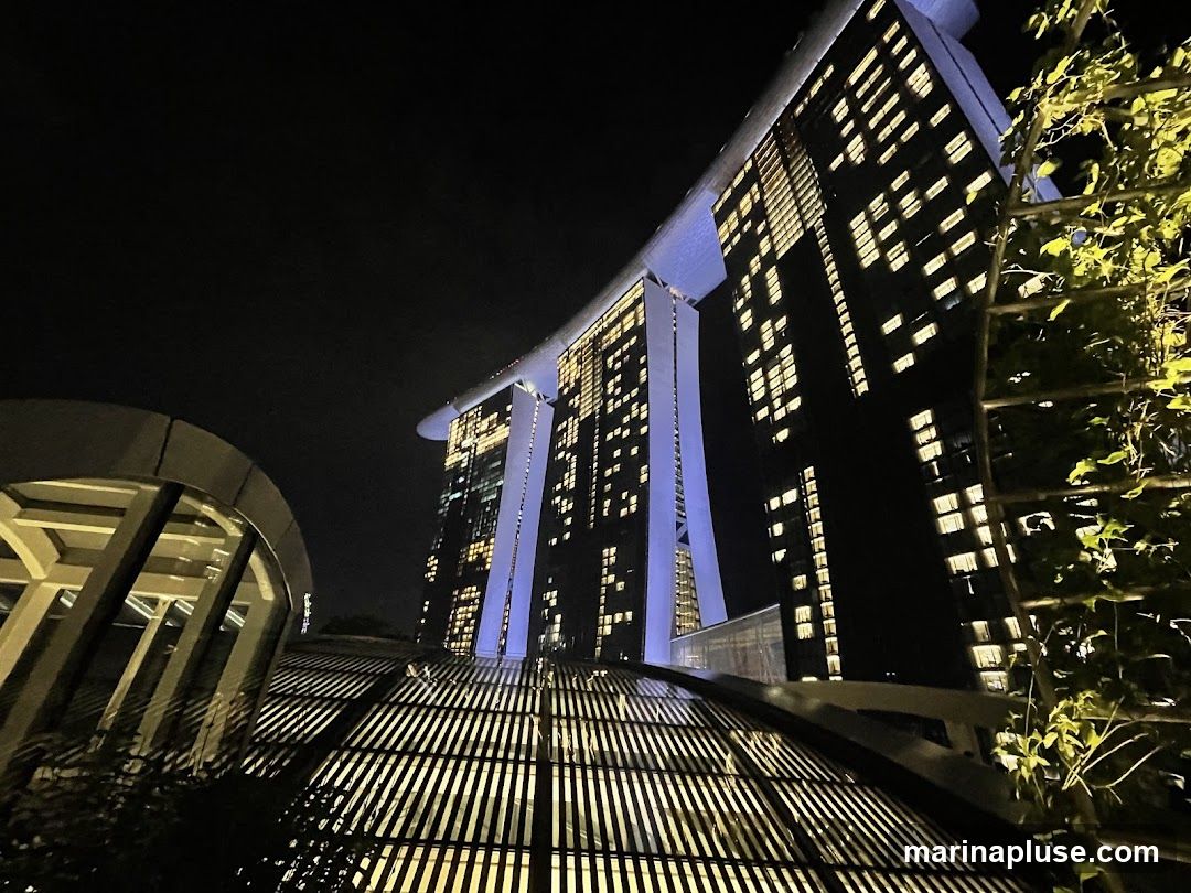A low-angle night view of the Marina Bay Sands hotel in Singapore. The iconic building is illuminated with many windows lit up, and blue lights accent its structure. In the foreground, a modern architectural element with a slatted roof casts shadows, and lush green foliage is visible on the right.