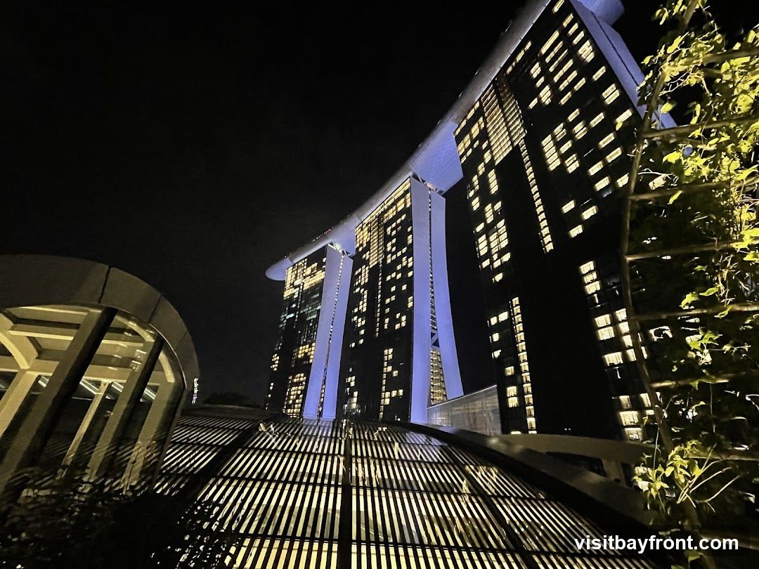 A low-angle night view of the Marina Bay Sands hotel in Singapore. The iconic building is illuminated with many windows lit up, and blue lights accent its structure. In the foreground, a modern architectural element with a slatted roof casts shadows, and lush green foliage is visible on the right.