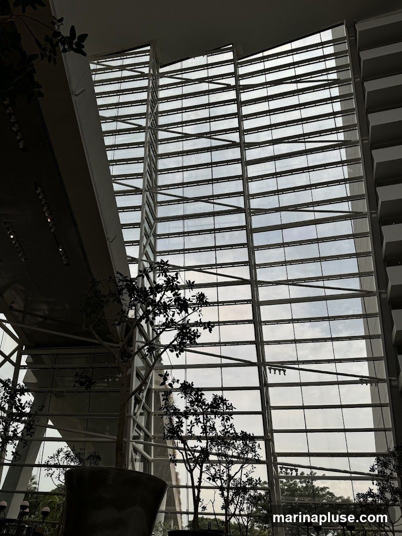The image shows the interior of a large, modern building with a soaring atrium. A massive glass facade dominates the right side of the frame, allowing natural light to filter in. The structure is characterized by its geometric lines and metal framework. In the foreground, silhouetted artificial trees in large planters add a touch of greenery to the minimalist aesthetic. The overall impression is one of spaciousness and contemporary design.