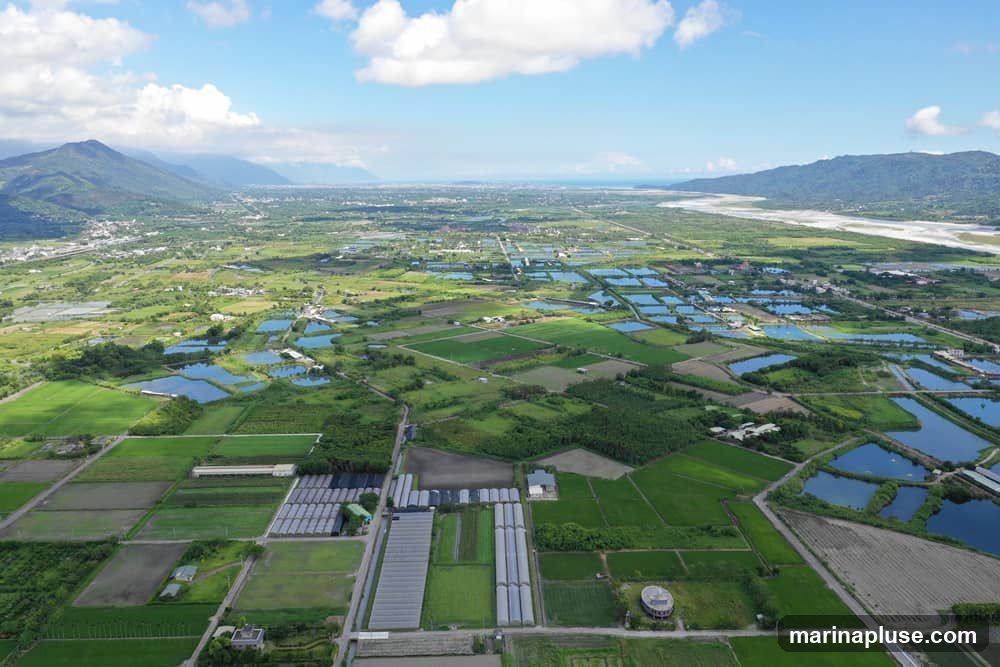 An aerial drone shot captures a vast agricultural landscape under a bright blue sky with scattered white clouds. The scene is dominated by numerous rectangular ponds, likely for aquaculture, interspersed with green fields and some greenhouses. In the distance, mountains frame the horizon, and a river or coastline is visible. The overall impression is one of rural productivity and natural beauty.