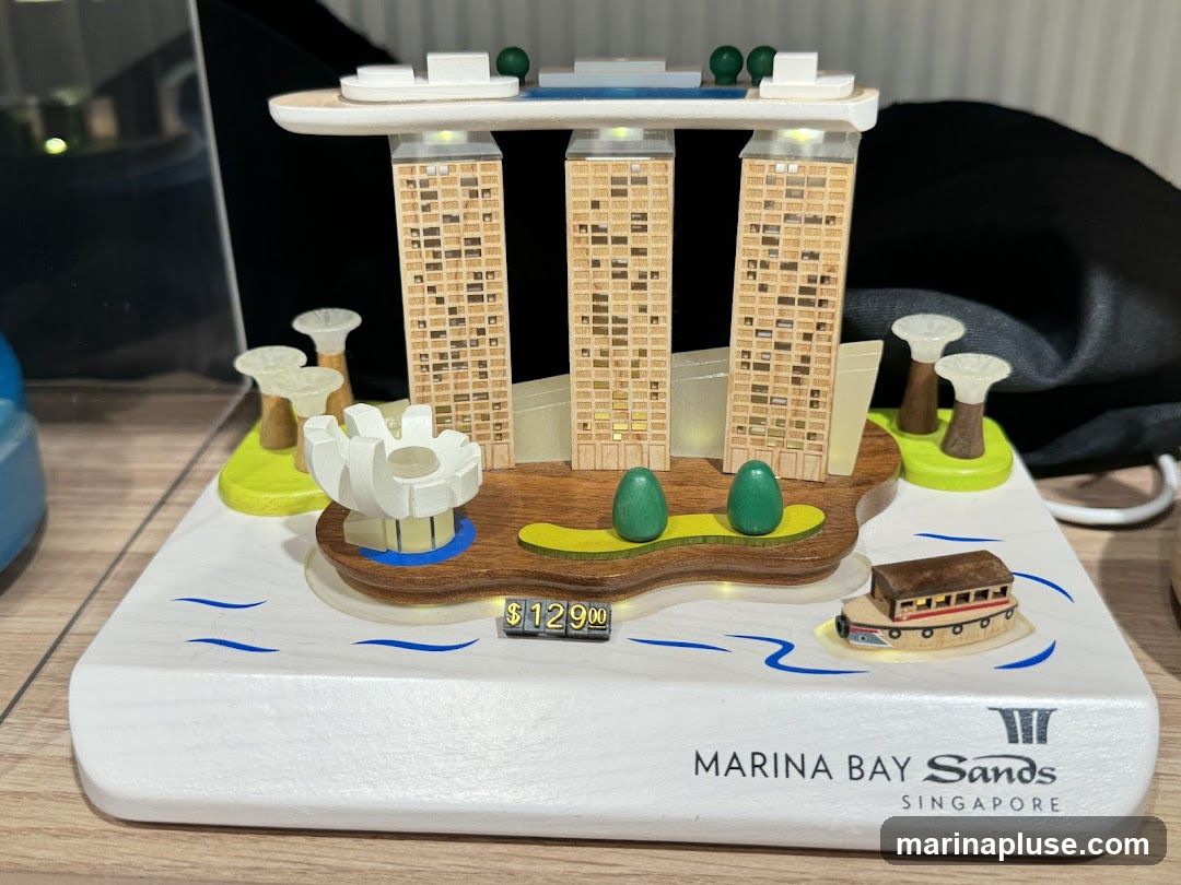 A miniature replica of the Marina Bay Sands hotel in Singapore is displayed on a white base. The replica features the iconic three towers connected by a sky park, with smaller decorative elements representing gardens and a boat. A price tag of $129.00 is visible below the replica. The base of the souvenir has the "Marina Bay Sands Singapore" logo printed on it.