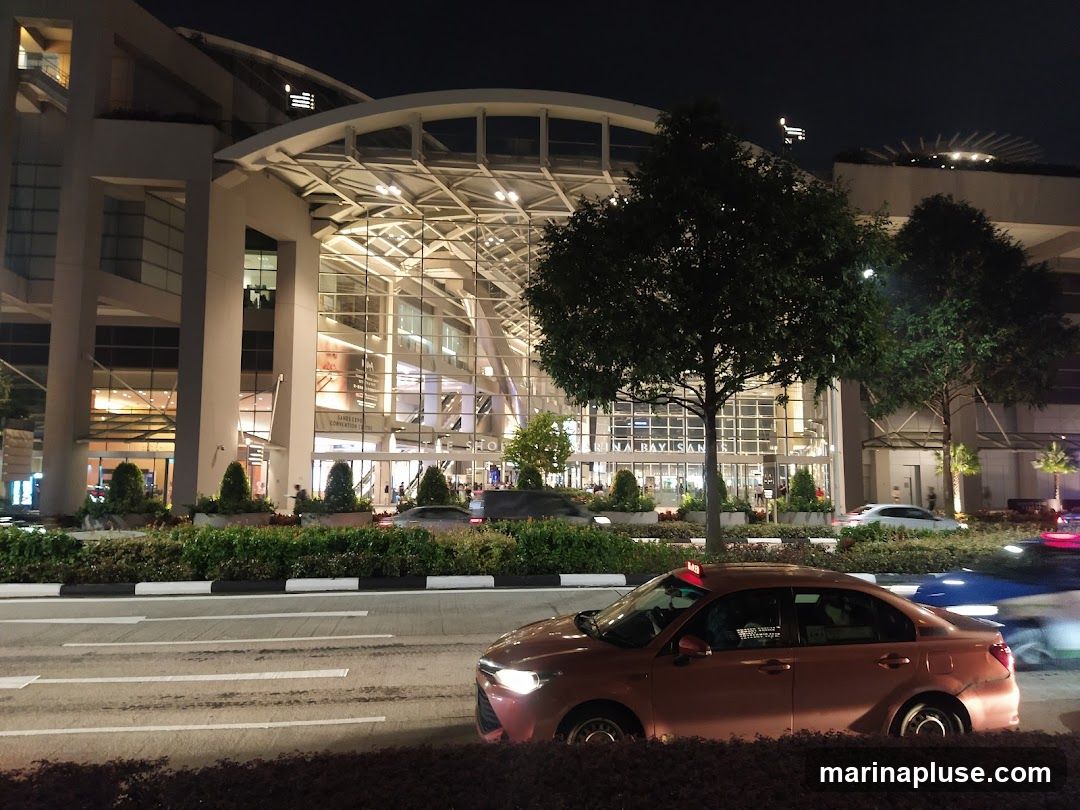 A wide shot of the Marina Bay Sands complex at night, with a street in the foreground. Cars are driving on the street, and the building's facade is illuminated. Trees and landscaping are visible in front of the building.