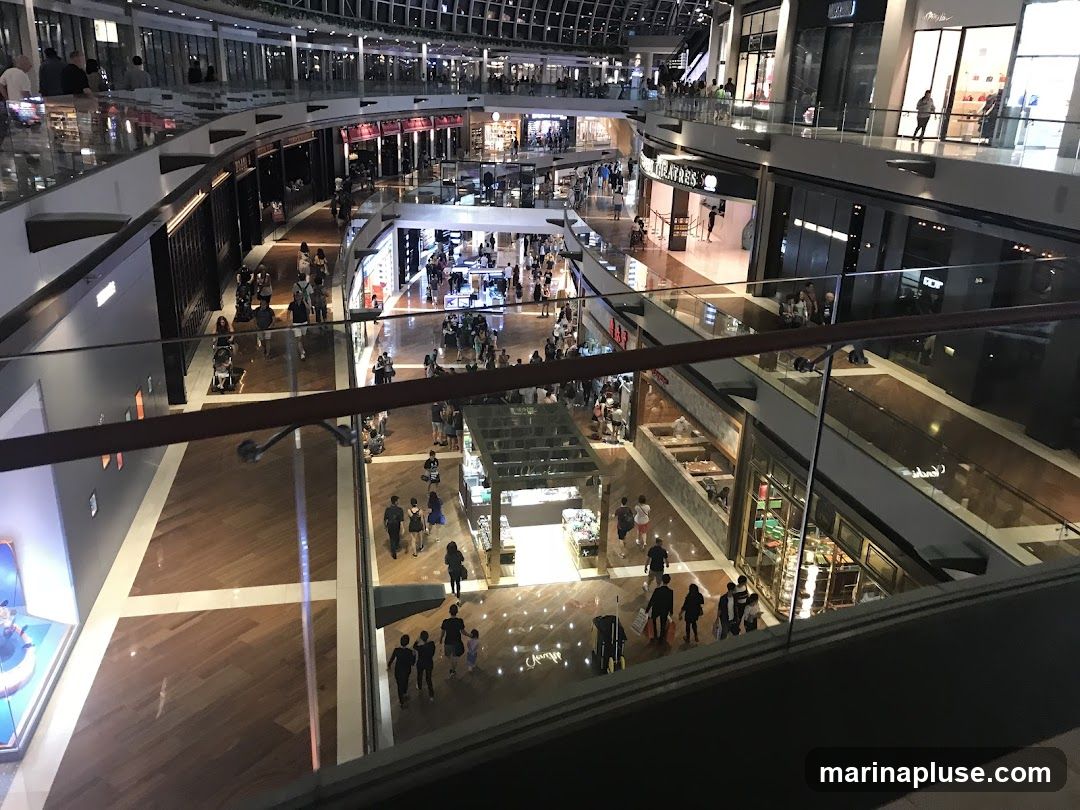 An overhead view of a bustling shopping mall with multiple levels. People are seen walking on the wooden floors and browsing in various shops. The architecture features curved walkways and glass railings, with shops lining the perimeter of each level. The mall is well-lit, with a modern and spacious design.