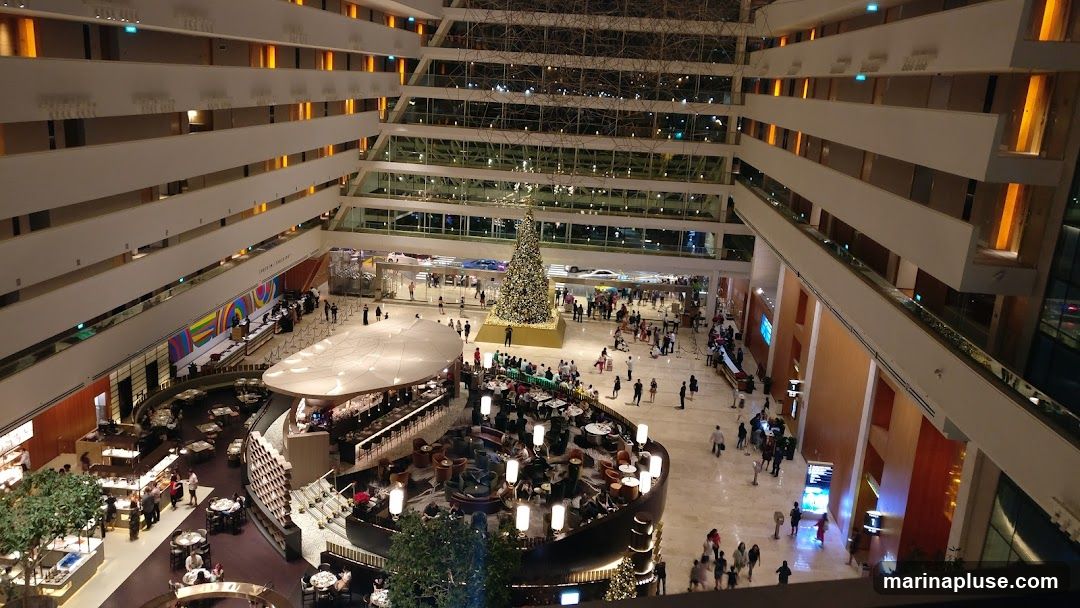 A wide shot of a hotel atrium, decorated for Christmas with a large, illuminated tree in the center of the lobby. People are seen milling about on the ground floor and on the various levels of the atrium. There are seating areas and a bar visible on the lower levels. The architecture features multiple tiers with balconies overlooking the central space.