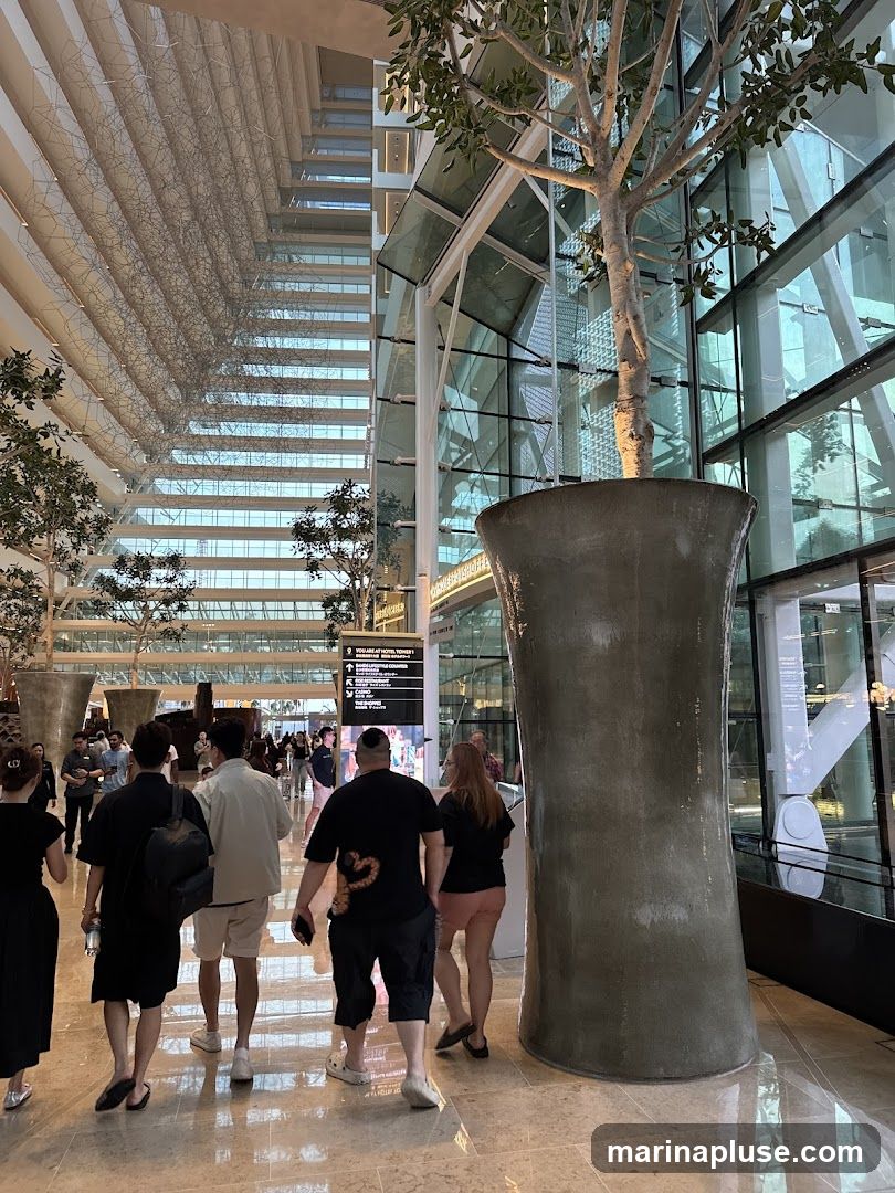 A wide shot of a modern indoor atrium with people walking. The architecture features a high ceiling with intricate geometric patterns and large glass windows. Several trees in large planters are placed throughout the space. The floor is polished marble, reflecting the light and architecture. A directional sign is visible in the background.