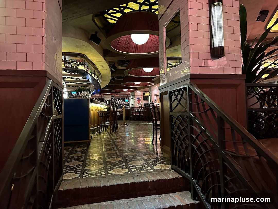 A view down a hallway in a restaurant, with decorative tiling on the floor and ceiling. Large pendant lights hang from the ceiling, and there are tables and chairs visible in the background. A bar area is also visible.