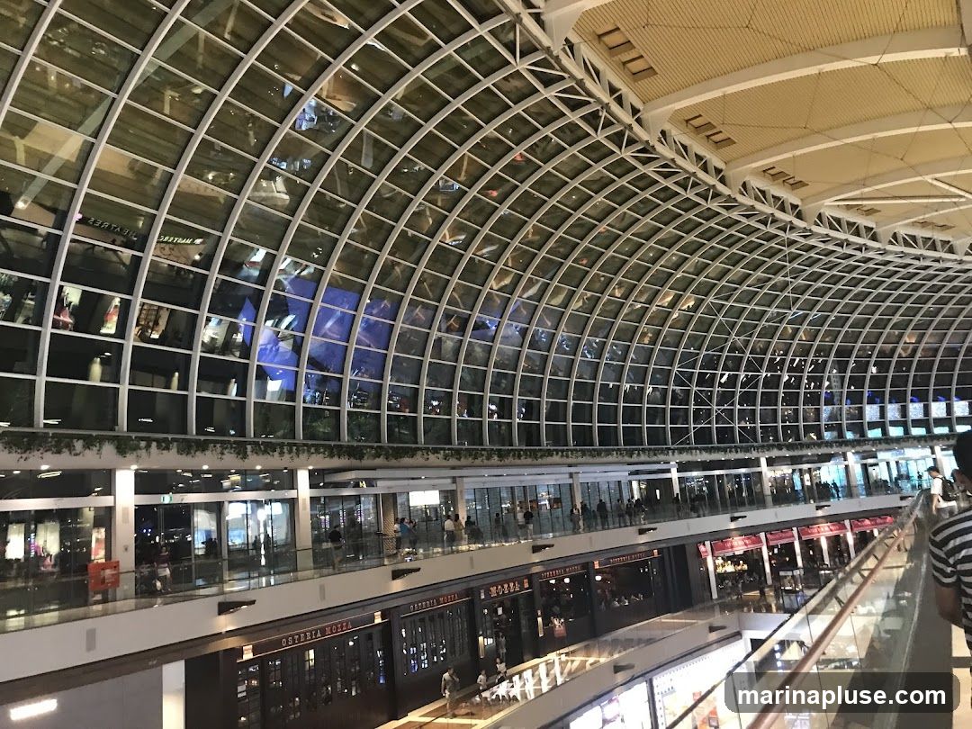 An interior view of a multi-level shopping mall with a distinctive arched glass ceiling. The architecture features a grid-like structure supporting the glass panels, reflecting the lights and activity within. Multiple levels of shops and walkways are visible, with people browsing and moving around. One shop prominently displays the name "OSTERIA MOZZA" on its facade. The overall impression is one of a spacious and contemporary commercial environment.