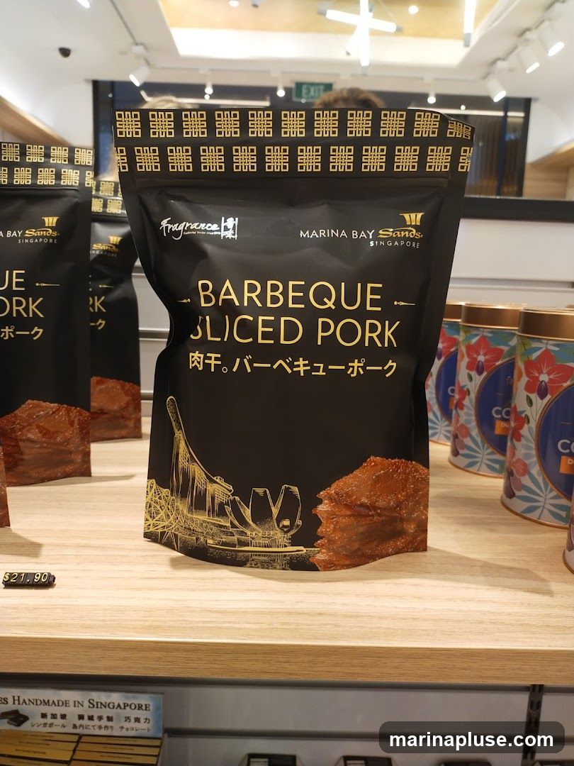 A close-up shot of a black retail package of barbeque sliced pork. The package features gold lettering and graphics, including the Marina Bay Sands Singapore logo and an illustration of the Marina Bay Sands complex. The product itself is visible on the packaging. A price tag of $21.90 is displayed below the package. Other similar packages and decorative tins are visible on the shelf.