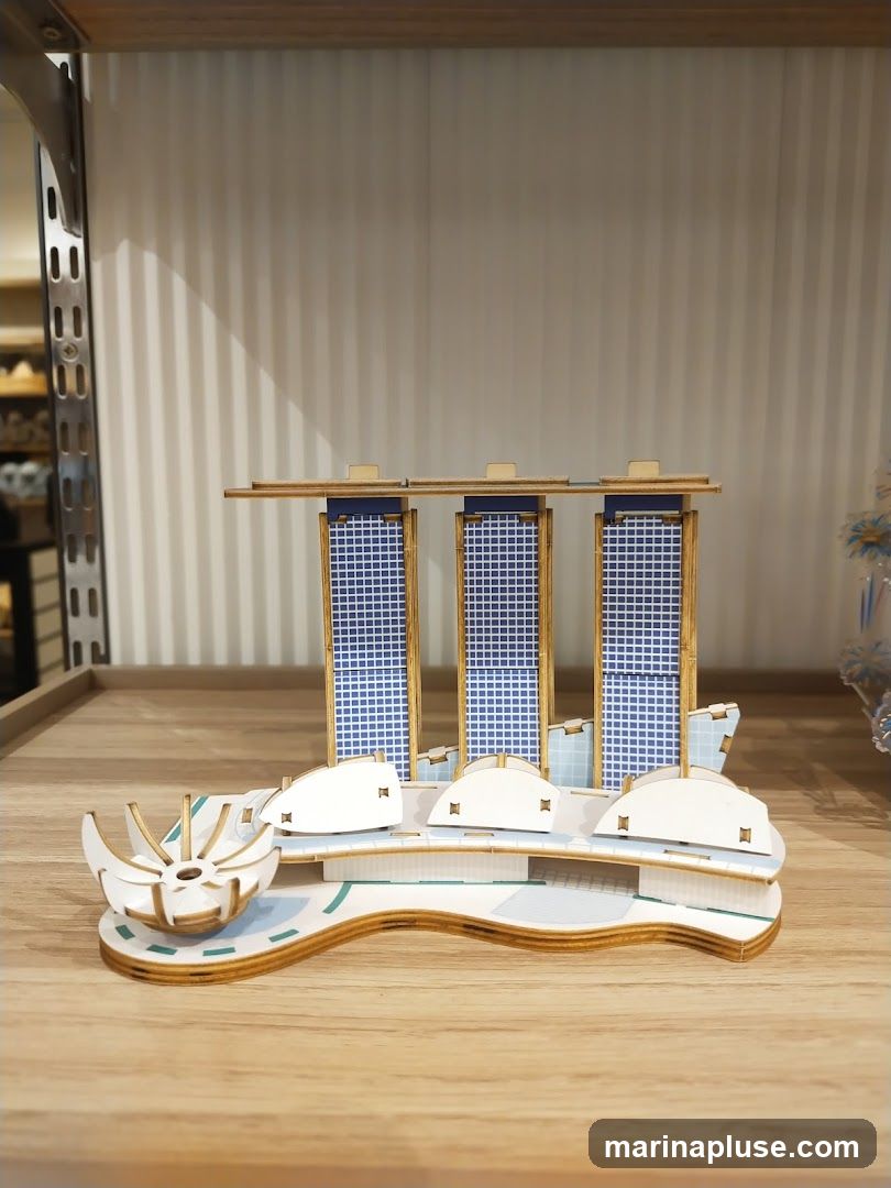 A 3D puzzle model of a building complex with three towers and a curved base is displayed on a wooden surface. The towers have blue checkered facades and wooden frames. The base features white architectural elements and a light blue pathway. In the background, a white corrugated wall is visible, and to the left, a metal shelf with items is partially seen.