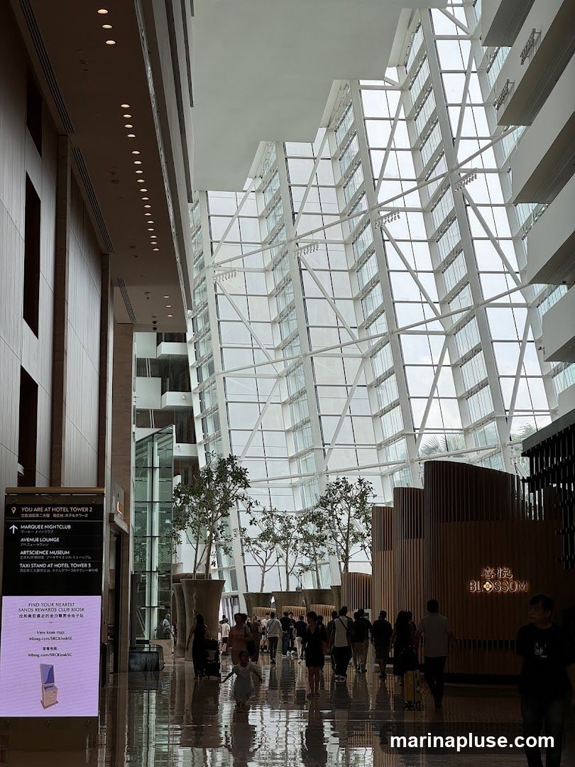 A wide shot of a modern hotel lobby with a striking glass ceiling and a wooden-paneled wall. People are walking through the spacious area, which features potted trees and a digital directory. A sign indicates directions to various hotel amenities, including a nightclub, lounge, museum, and taxi stand. The sign also promotes a Sands Rewards Club Kiosk. To the right, a section of the building is adorned with vertical wooden slats and a sign that reads "BLOSSOM" in English and Chinese characters.