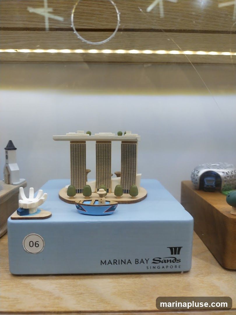 A miniature model of the Marina Bay Sands hotel in Singapore is displayed on a light blue box. The model includes the iconic three towers and a sky park. In front of the hotel model, there is a small blue boat with a figure inside. To the left of the hotel model, there is a small white building with a steeple. To the right, a silver structure resembling a tunnel entrance is visible. The box has the text "MARINA BAY Sands SINGAPORE" and a logo printed on it. A circular sticker with the number "06" is also present on the box. The display is lit from above and appears to be behind glass.