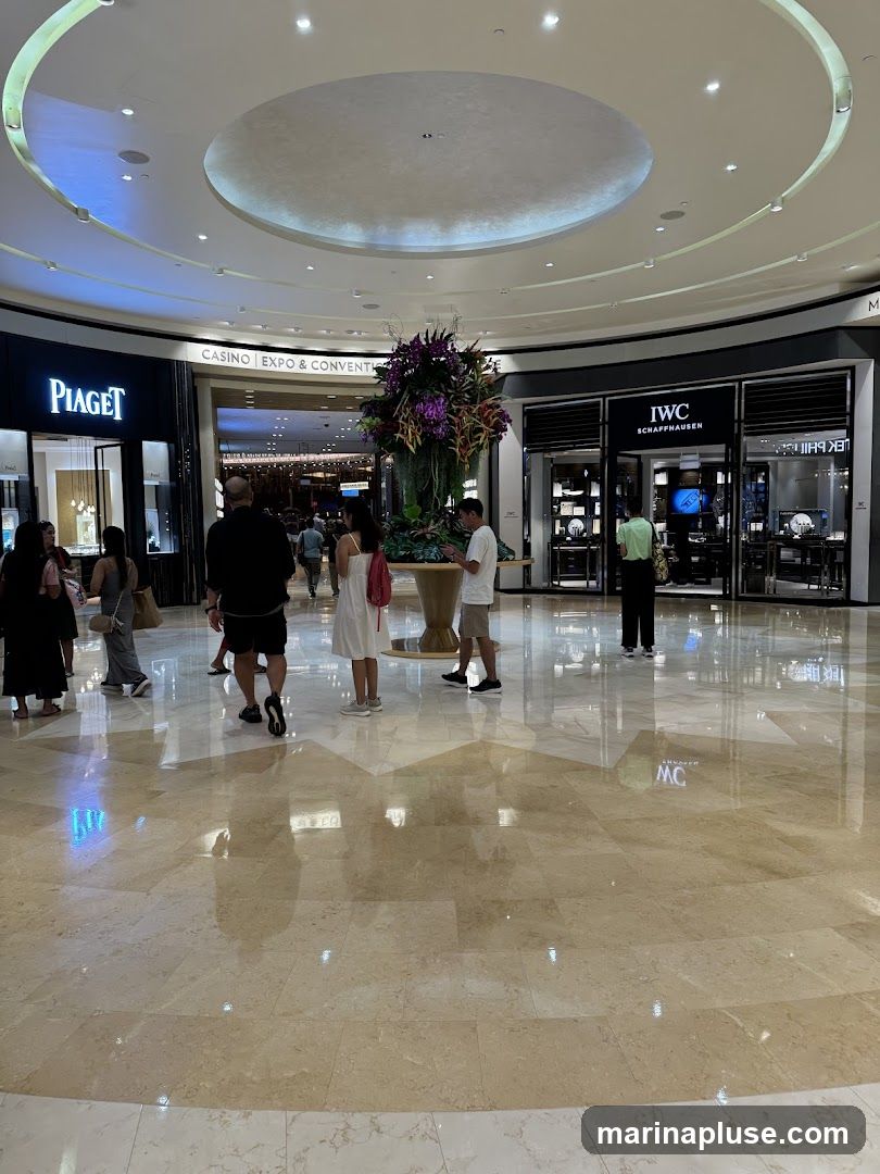 People walk through a brightly lit mall with polished marble floors. On the left is a Piaget store, and on the right is an IWC Schaffhausen store. In the center, a large floral arrangement sits in a planter. Beyond the stores, a sign indicates "CASINO EXPO & CONVENTION".