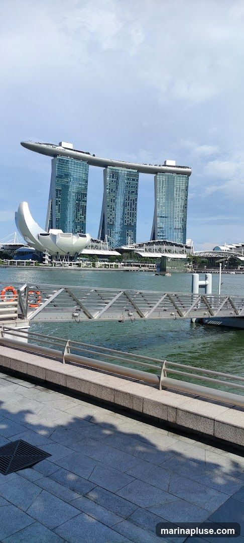 A wide shot of the Marina Bay Sands complex in Singapore, with its iconic three towers topped by a ship-like structure. The ArtScience Museum, with its lotus-like design, is visible in the foreground. A body of water separates the viewer from the buildings, and a metal walkway with railings is in the foreground. The sky is partly cloudy.