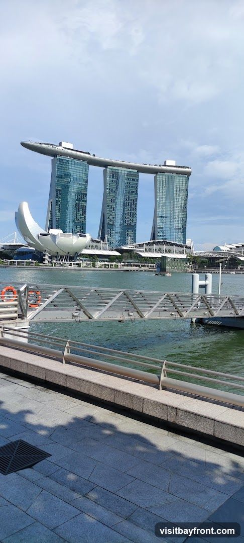 A wide shot of the Marina Bay Sands complex in Singapore, with its iconic three towers topped by a ship-like structure. The ArtScience Museum, with its lotus-like design, is visible in the foreground. A body of water separates the viewer from the buildings, and a metal walkway with railings is in the foreground. The sky is partly cloudy.