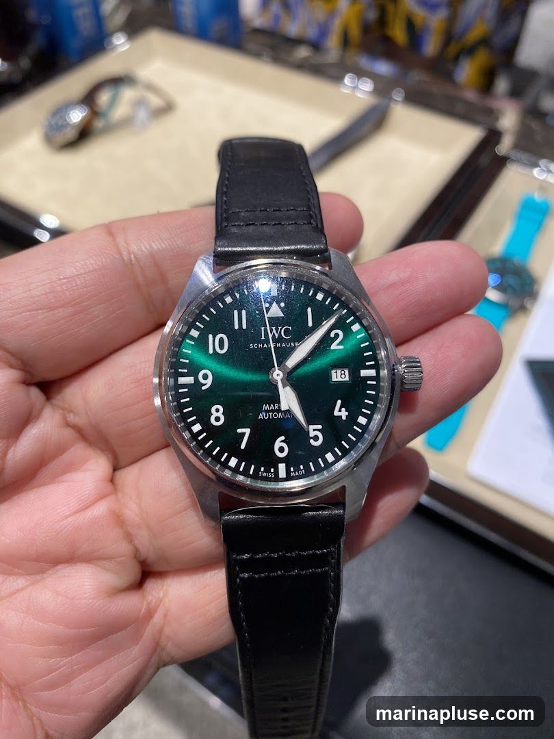 A close-up shot of a person holding an IWC Pilot's watch with a green dial and a black leather strap. The watch face displays Arabic numerals, a date window at 3 o'clock, and the IWC Schaffhausen logo. In the blurred background, other watches and display cases are visible.
