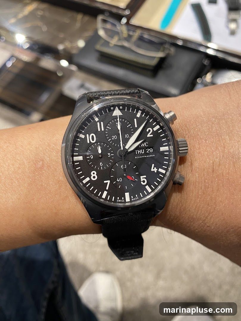 A close-up shot of a person's wrist wearing a black IWC Pilot's Watch Chronograph. The watch has a black dial with white Arabic numerals and sub-dials. The date window shows "THU 29". The watch is on a black leather strap. In the blurred background, eyeglasses and other items are visible on a surface.