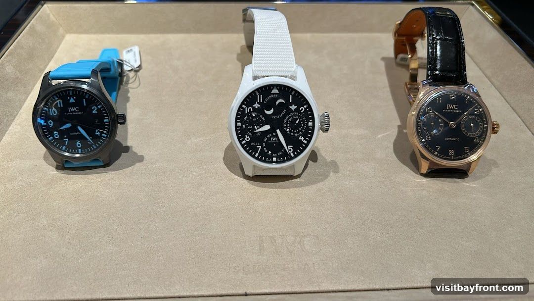 A display of three IWC watches on a textured surface. The watch on the left has a dark dial and a bright blue rubber strap. The middle watch has a white case and strap, with a complex dial showing multiple complications including a moon phase and perpetual calendar. The watch on the right has a rose gold case and a black alligator leather strap, with a dark dial and two sub-dials. The IWC logo is visible on the surface below the watches.