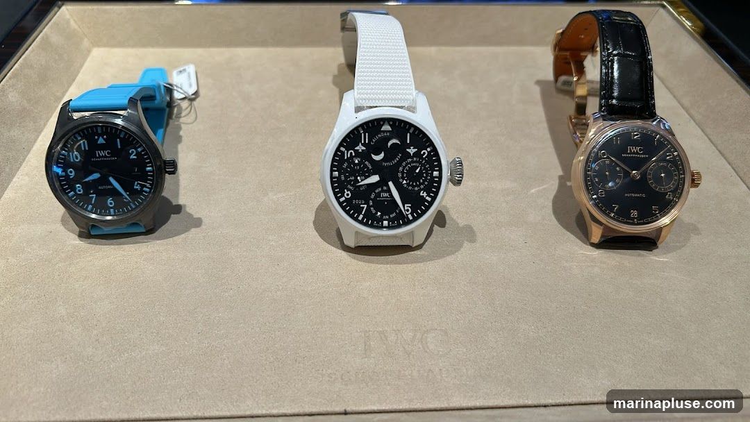 A display of three IWC watches on a textured surface. The watch on the left has a dark dial and a bright blue rubber strap. The middle watch has a white case and strap, with a complex dial showing multiple complications including a moon phase and perpetual calendar. The watch on the right has a rose gold case and a black alligator leather strap, with a dark dial and two sub-dials. The IWC logo is visible on the surface below the watches.