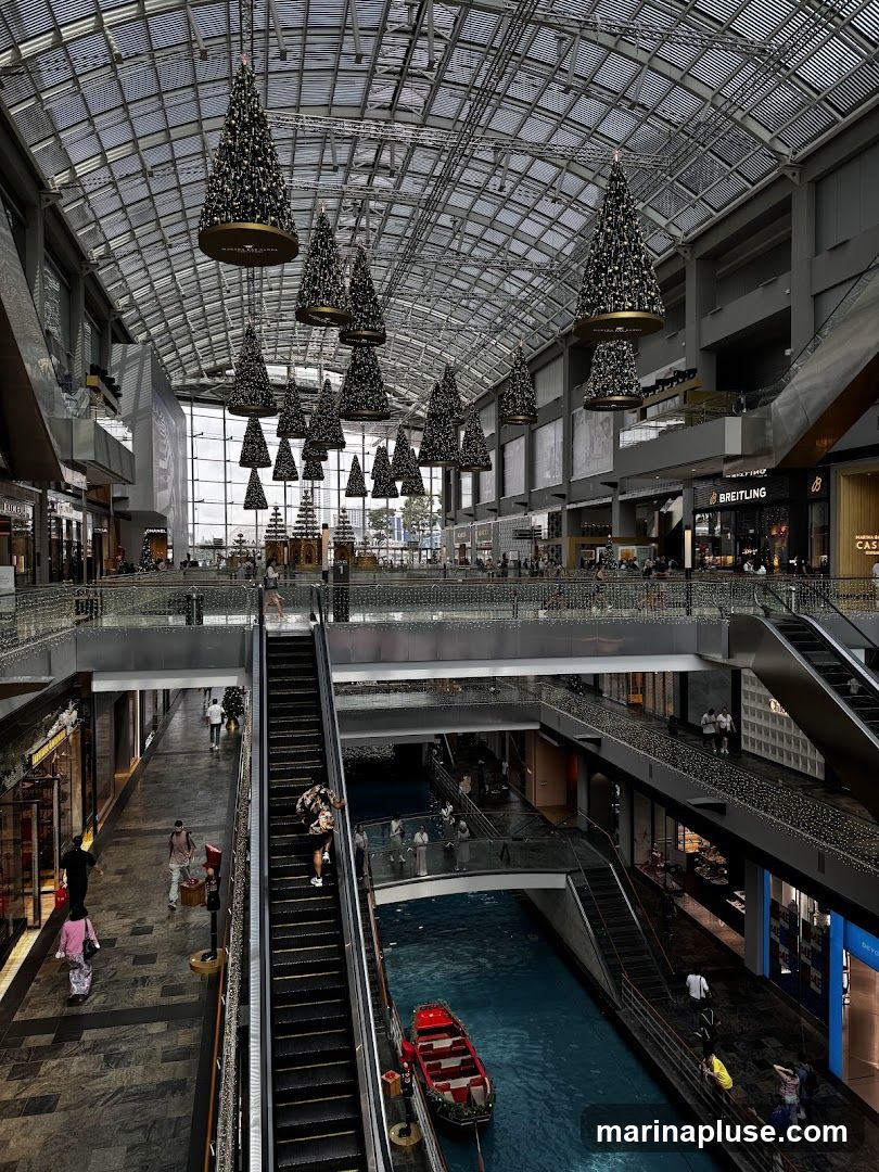 An indoor shopping mall with a glass ceiling, decorated with numerous Christmas trees hanging from above. Escalators and walkways are visible on multiple levels, with a canal running through the center of the mall, featuring a small red boat. People are seen shopping and walking around. Storefronts for various brands, including Breitling, are visible.