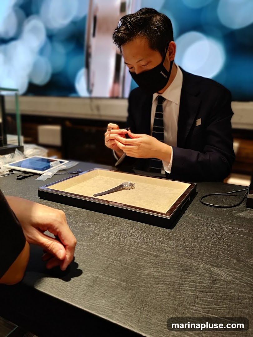 A man in a suit and mask is carefully examining a watch with tools. The watch is displayed on a tray on a dark textured table. Another person's hand is visible in the foreground. The background is blurred with bokeh lights.
