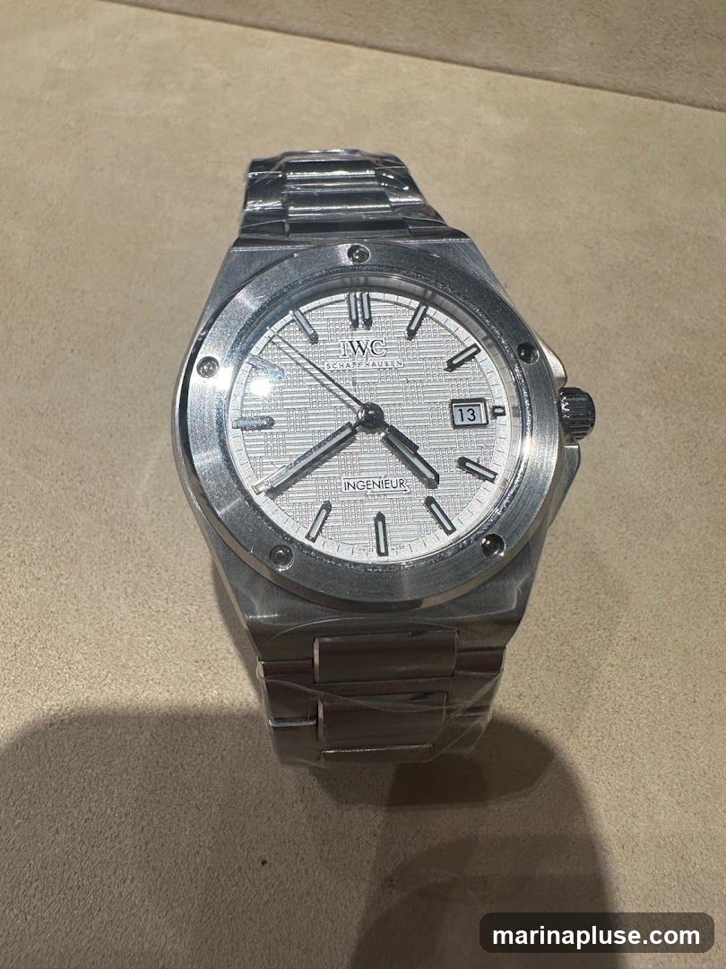 A close-up shot of a silver IWC Ingenieur watch with a white textured dial. The watch is covered in protective plastic film. The dial features silver hour markers, hands, and a date window at the 3 o'clock position. The IWC Schaffhausen logo is visible at the 12 o'clock position. The watch has a metal link bracelet.