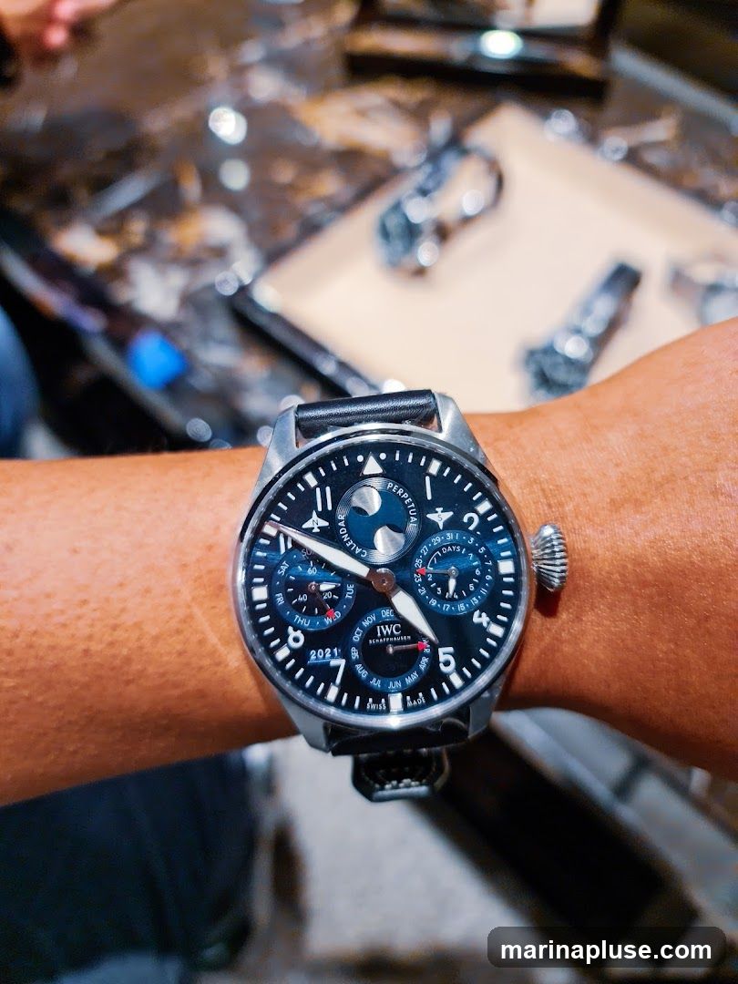 A close-up shot of a person's wrist wearing an IWC Schaffhausen Perpetual Calendar watch. The watch face is dark blue with silver hands and markers. It displays the date, day of the week, month, year, and a moon phase complication. The watch has a black leather strap. In the blurred background, other watches and a textured surface are visible.