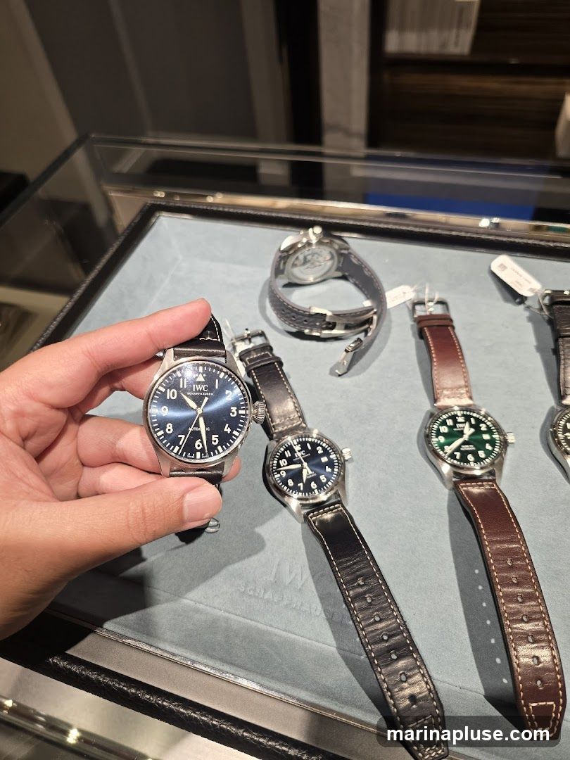 A hand holds a blue-faced IWC watch with a black leather strap. Several other watches with leather straps are displayed in a glass case. One watch has a green face, and another has a brown face. The watches are arranged on a grey velvet-like surface with the IWC logo embossed.