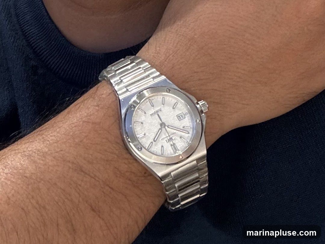 A close-up shot of a person's wrist wearing a silver metal watch. The watch has a textured white dial with Roman numerals and a date window showing '32'. The brand name 'IWC' is visible on the dial. The watch is attached to a metal link bracelet. The person is wearing a dark blue garment.