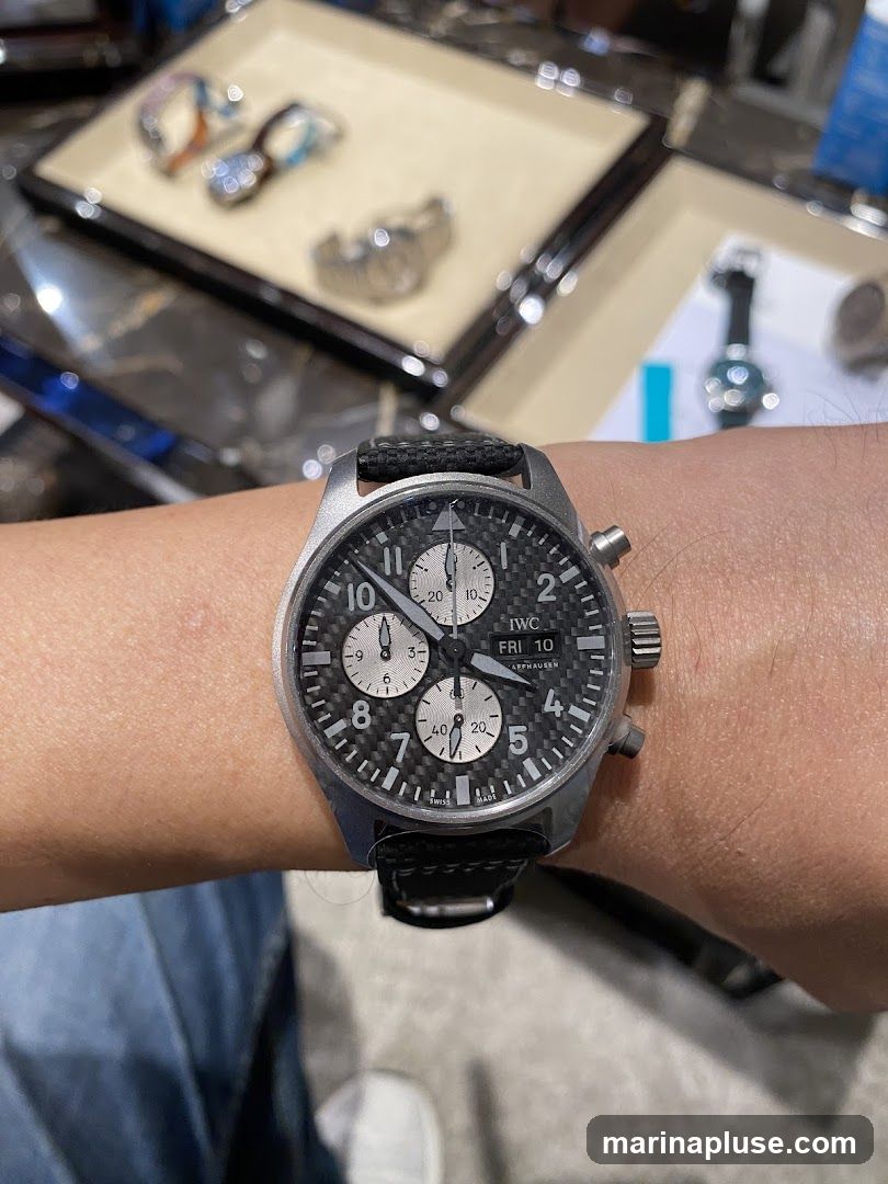 A close-up shot of a person's wrist wearing a chronograph watch with a black carbon fiber dial. The watch has silver hands and markers, with three sub-dials. The date window shows "FRI 10". In the blurred background, several other watches are displayed on trays.