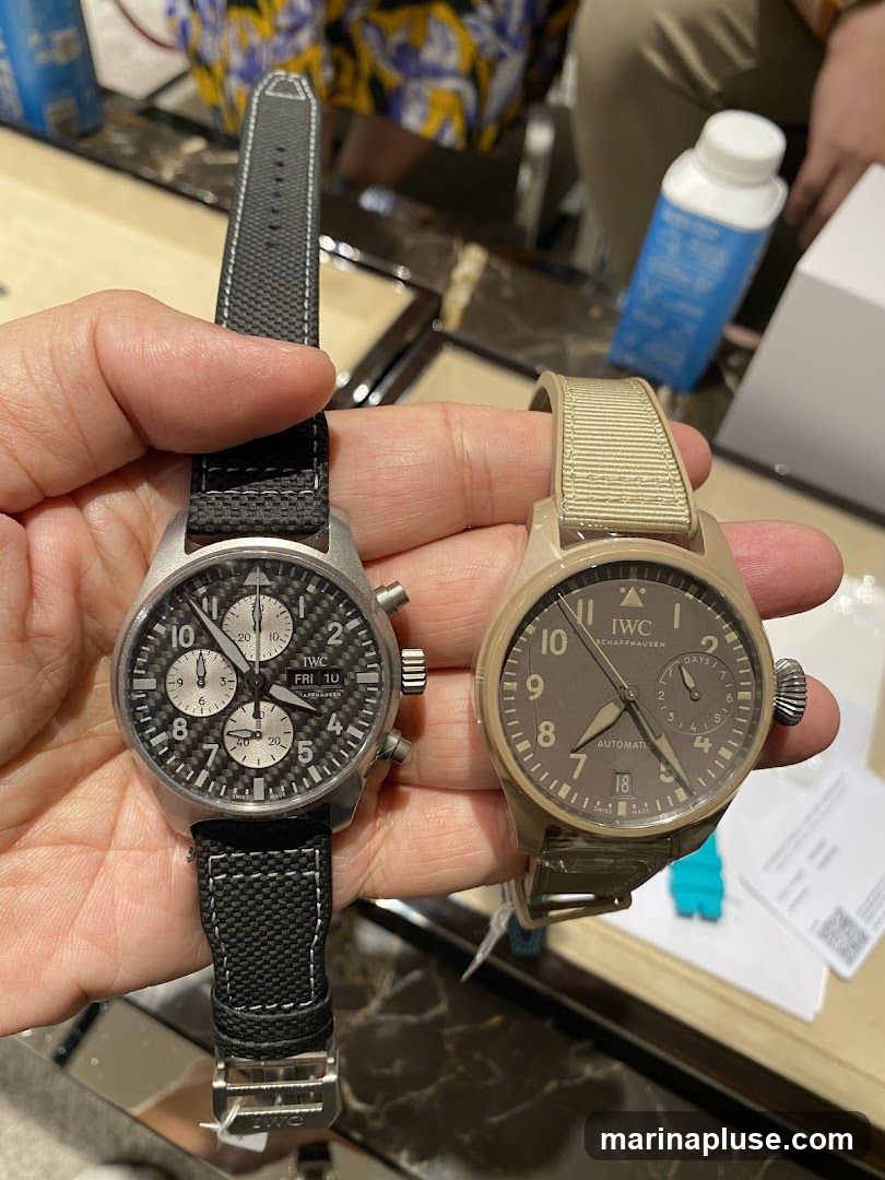 A person is holding two IWC watches. The watch on the left has a black strap and a carbon fiber dial with three subdials and a day/date complication. The watch on the right has a tan strap and a brown dial with a date window and a power reserve indicator. Both watches have the IWC Schaffhausen logo. In the background, there are display cases and a person wearing a colorful shirt.