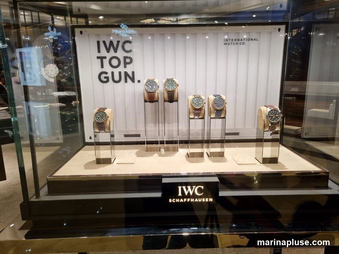 A display case featuring several IWC Top Gun watches. The watches are presented on individual stands within a glass enclosure. The background of the display has the text "IWC TOP GUN" and "INTERNATIONAL WATCH CO.". Below the display, the IWC Schaffhausen logo is visible.