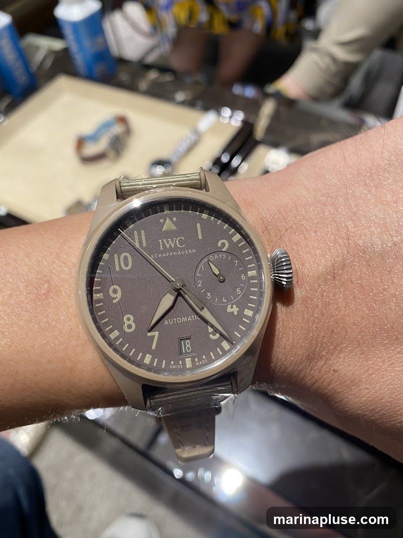 A close-up shot of a person's wrist wearing a IWC Pilot's Watch. The watch has a brown dial with Arabic numerals and a date window at the 3 o'clock position. The watch band is made of light brown leather. In the background, there are blurred images of people and display cases, suggesting a retail environment.