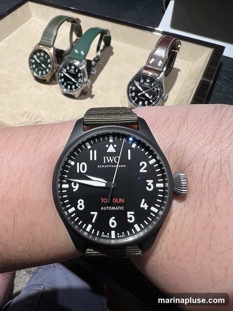 A close-up of a person wearing a black IWC Pilot's Watch with a green fabric strap. The watch face is black with white Arabic numerals and hands. Text on the dial reads "IWC SCHAFFHAUSEN", "TO GUN", and "AUTOMATIC". In the background, three other IWC watches are displayed in a tray, two with green straps and one with a brown leather strap.