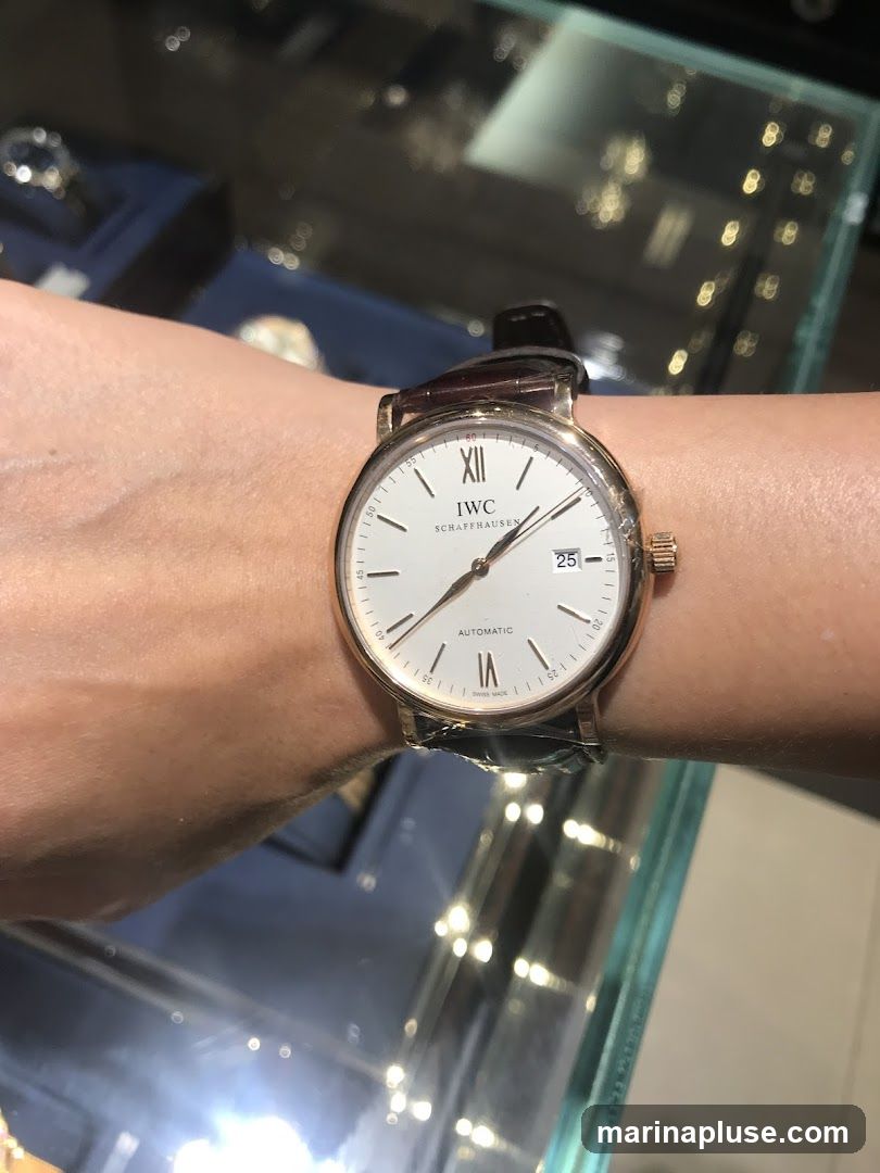 A close-up shot of a person wearing a white-faced IWC Schaffhausen automatic watch with a brown leather strap. The watch face displays Roman numerals for the hour markers, with XII at the top. The brand name "IWC SCHAFFHAUSEN" is visible below the XII. The date "25" is shown in a small window at the 3 o'clock position. The word "AUTOMATIC" is printed below the center hands. The background appears to be a display case in a jewelry or watch store, with blurred lights and reflections.