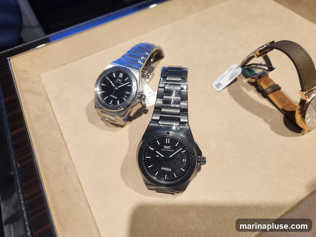 Three IWC watches are displayed on a beige surface. The watch in the foreground is dark gray with a black dial and a metal band. The watch to its left is silver with a black dial and a metal band. The watch on the right has a rose gold case and a brown leather strap. All watches have visible branding.